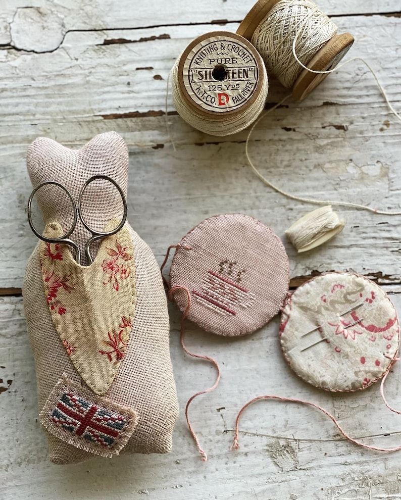 Puede incluir: Un coj&iacute;n de alfileres de tela beige con un dise&ntilde;o floral y un peque&ntilde;o parche de la Union Jack. El coj&iacute;n de alfileres est&aacute; lleno de agujas e hilo. Tambi&eacute;n hay dos carretes de hilo, uno con una etiqueta que dice "Knitting & Crochet Pure Sixteen 125 Yds Fall River K.T.Co."