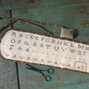 May include: A brown and white needlepoint alphabet sampler with a brown ribbon loop. The sampler is stitched with the letters of the alphabet in a simple font. The sampler is framed with a brown border and has a small brown and white needlepoint design at the bottom.