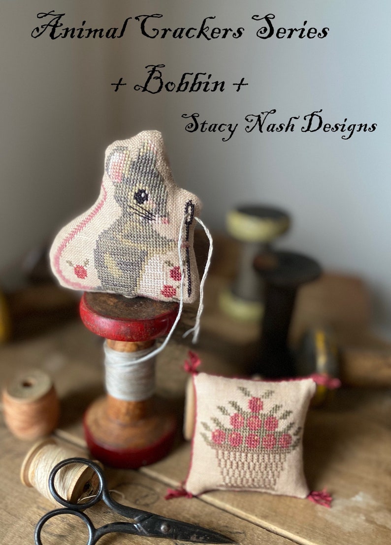 May include: A cross-stitch pattern of a mouse holding a needle and thread, with a basket of red berries. The pattern is on a small cushion, sitting on a wooden bobbin with a red top. The bobbin is on a wooden surface with other bobbins of thread and a pair of scissors. The text "Animal Crackers Series + Bobbin + Stacy Nash Designs" is at the top of the image.