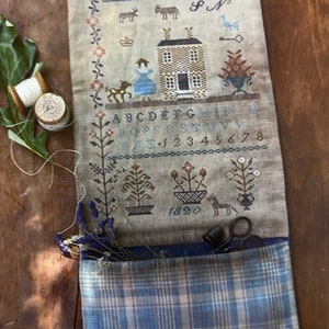 May include: A vintage-style embroidered wall hanging with the text "She seeketh wool and flax and worketh willingly with her hands." The design includes a house, figures, and floral motifs. A blue plaid pocket holds scissors and dried flowers.