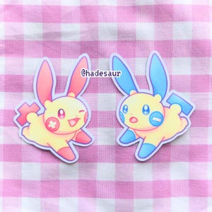 May include: Two yellow cartoon character stickers with blue and pink accents. The characters are wearing a plus and minus sign on their chests. The stickers are on a pink and white gingham background. @hadesaur