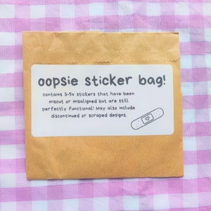 Puede incluir: Un sobre de papel marrón con una etiqueta blanca que dice "oopsie sticker bag! contains 3-5x stickers that have been miscut or misaligned but are still perfectly functional! May also include discontinued or scraped designs." Una ilustración en blanco y negro de una venda con un corazón en ella está en la etiqueta.