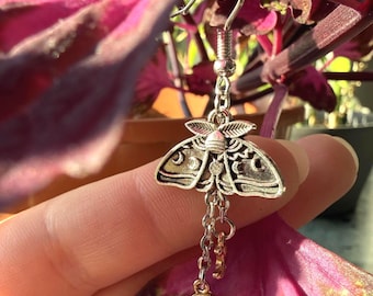 Puede incluir: Pendiente plateado con forma de polilla con intrincados detalles en las alas. El pendiente cuelga de una cadena con una pequeña estrella y un dije de media luna. El fondo presenta un follaje rosa y verde vibrante.