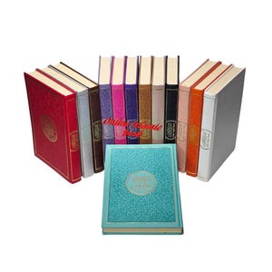 Op de afbeelding: Een verzameling hardcover boeken in verschillende kleuren, waaronder rood, wit, bruin, roze en blauwgroen. De boeken zijn in een halve cirkel gerangschikt, met één blauwgroen boek plat in het midden. De ruggen en kaften hebben gouden accenten.