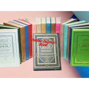 May include: A collection of hardcover books titled "The Noble Qur'an" in various colors, including white, green, blue, and gold. The text "Transliteration in Roman Script" is visible on the covers.