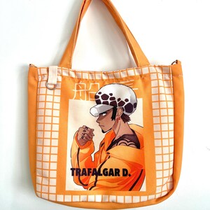 Peut inclure: Sac fourre-tout orange avec un motif quadrillé blanc et des poignées orange. Il présente une illustration d'un personnage en veste orange et chapeau blanc, avec le texte "TRAFALGAR D." en dessous.