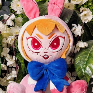 May include: A plush doll in the shape of a bunny girl, featuring pink and white fabric with stitched details. The doll has a blue bow, orange hair, and large red eyes. The bunny ears are pink and stitched.