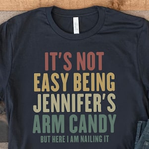 May include: A navy blue t-shirt with the text "It's Not Easy Being Jennifer's Arm Candy But Here I Am Nailing It" in retro colors. The shirt is paired with blue jeans and white sneakers.