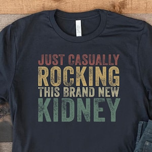 May include: A dark blue t-shirt with the text "Just Casually Rocking This Brand New Kidney" in a vintage style font. The shirt is laid out on a wooden surface with a pair of blue jeans and white trainers.
