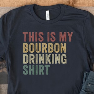 以下が含まれることがあります： ヴィンテージスタイルのフォントで「This is my Bourbon Drinking Shirt」と書かれた黒いTシャツ。Tシャツは、白いスニーカーと青いジーンズと一緒に木製の表面に置かれています。
