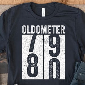 Op de afbeelding: Een marineblauw t-shirt met de tekst "OLDOMETER" boven de cijfers "79 80" in witte letters. Het shirt wordt gecombineerd met een blauwe jeans en witte sneakers.