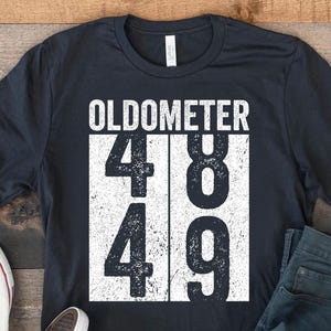 May include: A black t-shirt with the word "OLDOMETER" above the numbers 48 and 49 in a white rectangular design. The shirt is paired with blue jeans and white canvas sneakers.