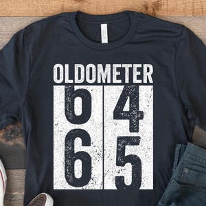 May include: A black t-shirt with the word "OLDOMETER" above the numbers 64 and 65. A pair of blue jeans and white sneakers are next to the shirt on a wooden surface.