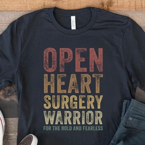 May include: A black t-shirt featuring a vintage-style graphic that reads "Open Heart Surgery Warrior For The Bold And Fearless". The shirt is styled with blue jeans and white trainers.