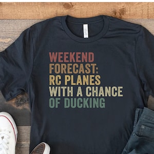 May include: A black t-shirt with a vintage-style graphic that reads "Weekend Forecast: RC Planes with a chance of Ducking". The shirt is laid out on a wooden surface with a pair of white sneakers and blue jeans.