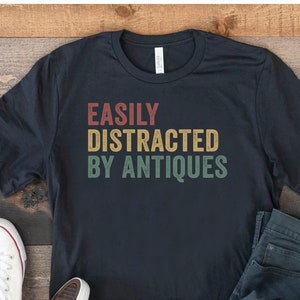 May include: A black t-shirt with the text "EASILY DISTRACTED BY ANTIQUES" in a vintage style font with yellow, red, and green colors.