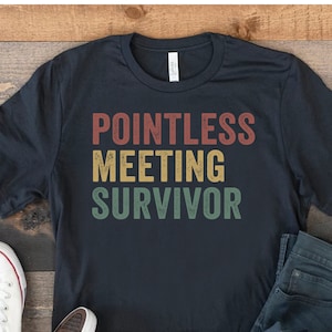 May include: A black t-shirt with the text "Pointless Meeting Survivor" in a vintage style font with red, yellow, and green colors. The shirt is laid out on a wooden surface with a pair of white sneakers and blue jeans.
