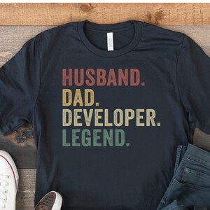 May include: A navy blue t-shirt with the words "HUSBAND. DAD. DEVELOPER. LEGEND." in a vintage style font. The shirt is paired with blue jeans and white canvas sneakers on a wooden surface.
