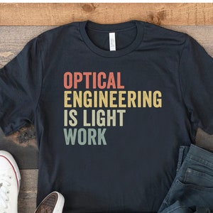 May include: Dark gray t-shirt with the text "OPTICAL ENGINEERING IS LIGHT WORK" in a retro color scheme. The shirt is paired with blue jeans and white canvas sneakers on a wooden surface.