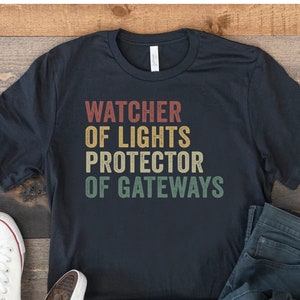 May include: A black t-shirt with a vintage-style graphic that reads "Watcher of Lights Protector of Gateways". The shirt is laid out on a wooden surface with a pair of white trainers and blue jeans.