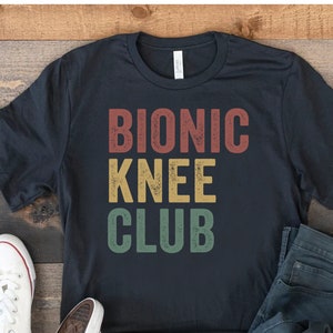 May include: A black t-shirt with the text "Bionic Knee Club" in a vintage style font with red, yellow, and green colours. The shirt is laid out on a wooden surface with a pair of white trainers and blue jeans.