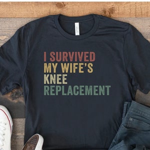May include: A black t-shirt with the text "I survived my wife's knee replacement" in a vintage style font. The shirt is laid out on a wooden surface with a pair of white sneakers and blue jeans.