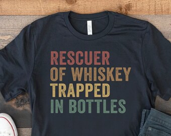 May include: A black t-shirt with a vintage-style graphic that reads "RESCUER OF WHISKEY TRAPPED IN BOTTLES". The shirt is laid out on a wooden surface with a pair of white sneakers and blue jeans.