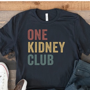May include: A black t-shirt with the text "ONE KIDNEY CLUB" in a vintage style font. The text is in red, yellow, and green.