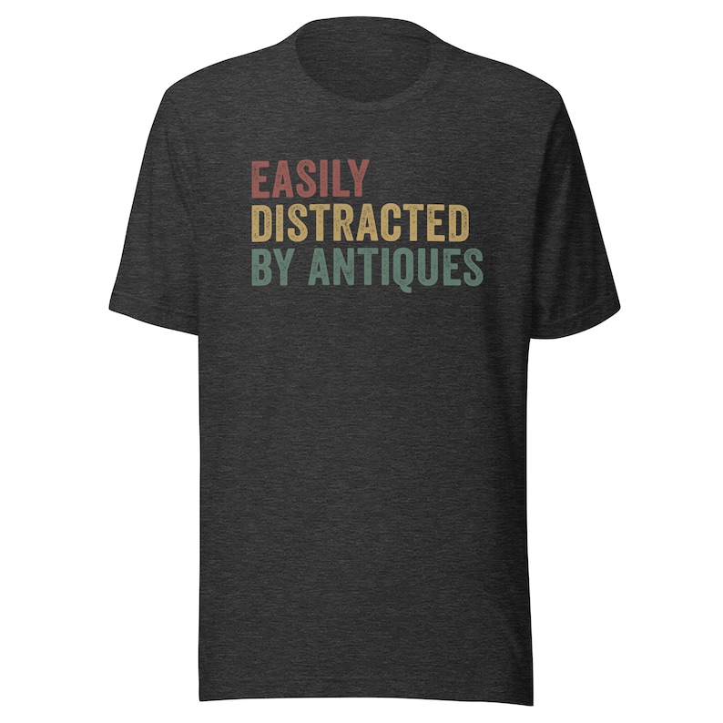 May include: A dark heather grey t-shirt with the text "EASILY DISTRACTED BY ANTIQUES" printed in a vintage style font with red, yellow, and green colors.