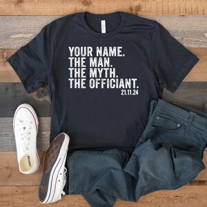 May include: Black t-shirt with white text that reads "YOUR NAME. THE MAN. THE MYTH. THE OFFICIANT. 21.11.24". The shirt is laid out on a wooden surface with a pair of white sneakers and blue jeans.