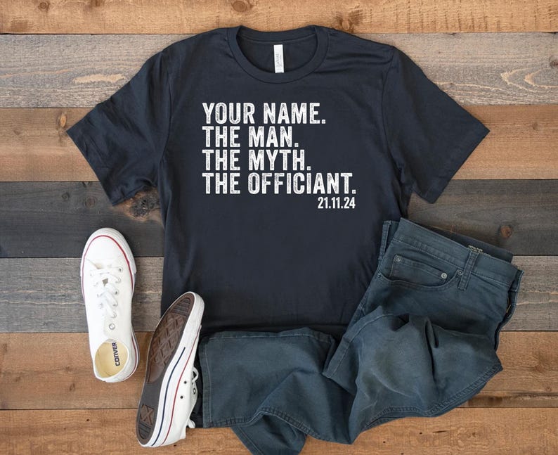 May include: Black t-shirt with white text that reads "YOUR NAME. THE MAN. THE MYTH. THE OFFICIANT. 21.11.24". The shirt is laid out on a wooden surface with a pair of white sneakers and blue jeans.
