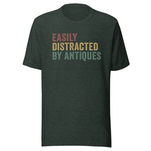 May include: A dark green t-shirt with the text "EASILY DISTRACTED BY ANTIQUES" in a vintage style font with red, yellow, and blue colors.