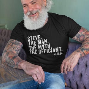 May include: A man wearing a black t-shirt with white text that reads "STEVE. THE MAN. THE MYTH. THE OFFICIANT. 21.11.24". He has a long white beard and is sitting on a purple couch.