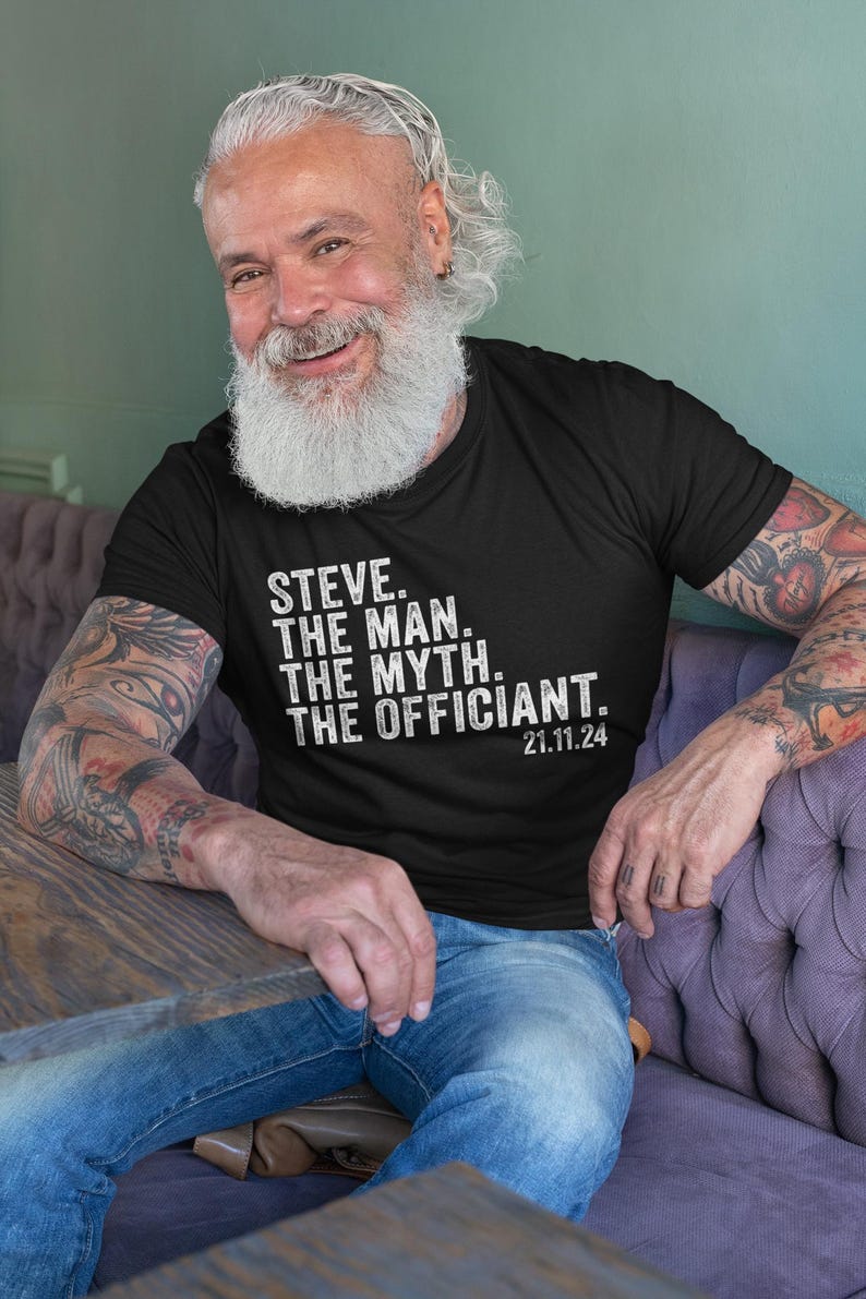 May include: A man wearing a black t-shirt with white text that reads "STEVE. THE MAN. THE MYTH. THE OFFICIANT. 21.11.24". He has a long white beard and is sitting on a purple couch.