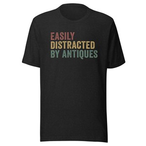 May include: Black t-shirt with the text "EASILY DISTRACTED BY ANTIQUES" in a vintage style font with red, yellow, and green colors.