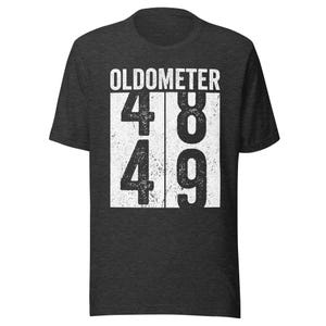 May include: Dark gray t-shirt with the word "OLDOMETER" above the numbers 48 and 49 in a white, distressed font. The shirt is a crew neck style.