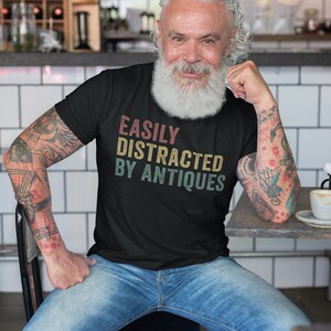 May include: A man wearing a black t-shirt with the text "EASILY DISTRACTED BY ANTIQUES" in a vintage style font. He is sitting on a chair with his arms resting on a table.