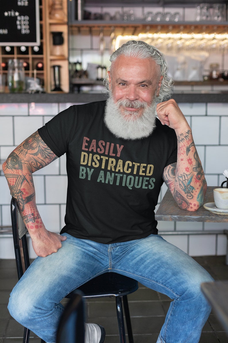 May include: A man wearing a black t-shirt with the text "EASILY DISTRACTED BY ANTIQUES" in a vintage style font. He is sitting on a chair with his arms resting on a table.