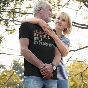 May include: A couple in a park, the man is wearing a black t-shirt with the text "I survived my wife's knee replacement" in a vintage style font. The couple is smiling and looking at each other.