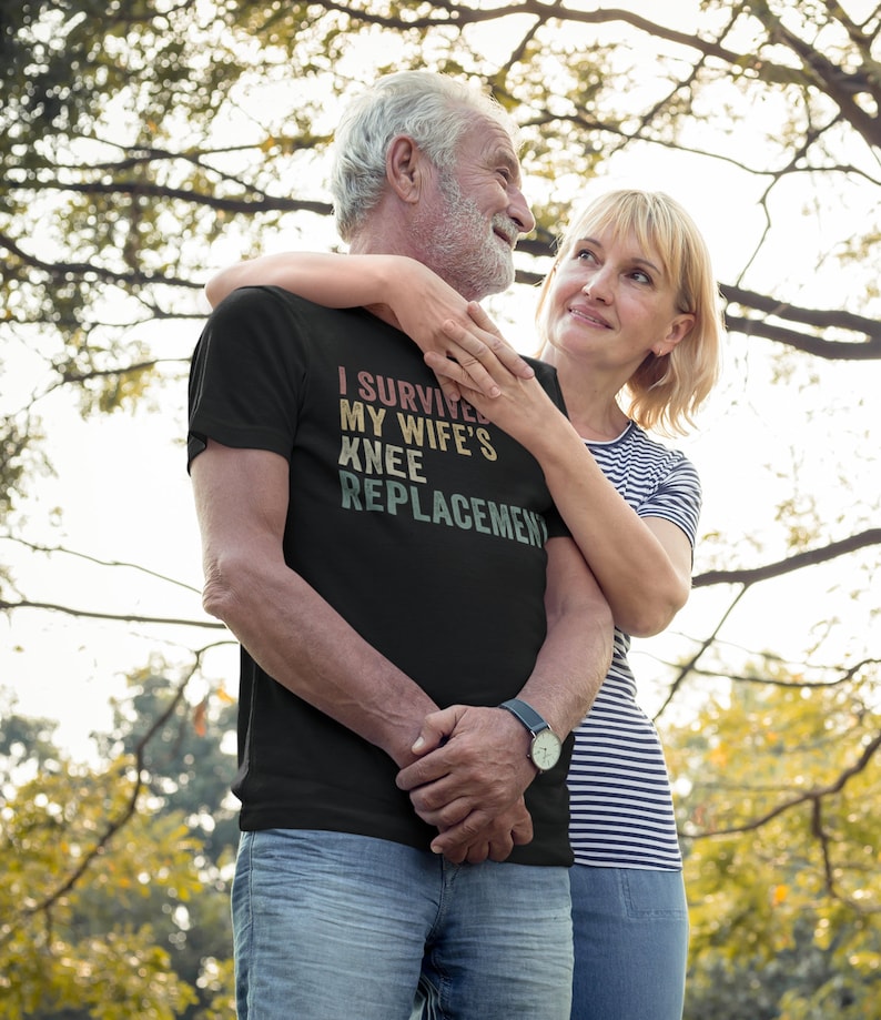 May include: A couple in a park, the man is wearing a black t-shirt with the text "I survived my wife's knee replacement" in a vintage style font. The couple is smiling and looking at each other.