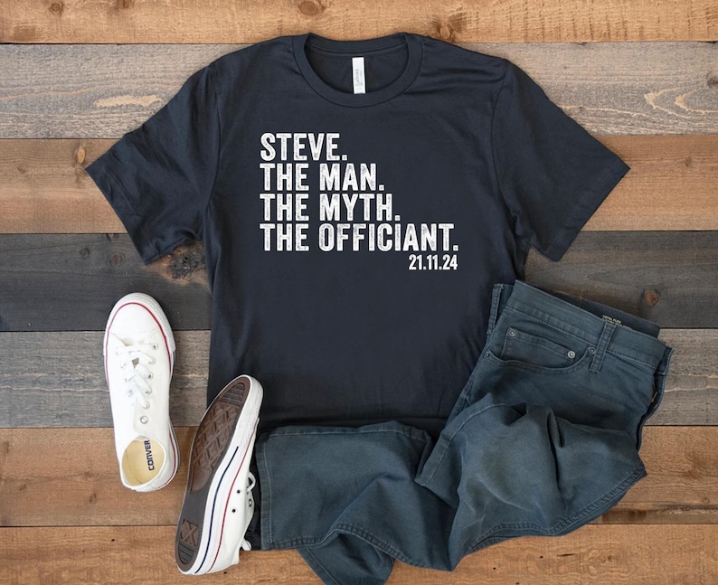May include: A black t-shirt with white text that reads "STEVE. THE MAN. THE MYTH. THE OFFICIANT. 21.11.24". The shirt is laid out on a wooden surface with a pair of white sneakers and blue jeans.