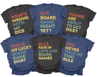 May include: Six t-shirts in various colors, each with a different board game-themed phrase in colorful text. Phrases include "What's wrong with my dice" and "Is it board game night yet?" The shirts are casual wear.