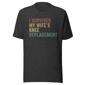 May include: A dark heather grey t-shirt with the text "I survived my wife's knee replacement" in a vintage style font.