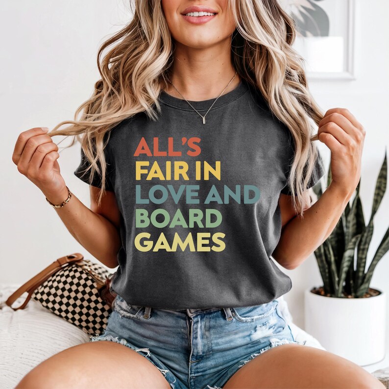 May include: Dark gray t-shirt with the text "ALL'S FAIR IN LOVE AND BOARD GAMES" in colorful letters. The shirt is a crew neck and has short sleeves. The shirt is worn by a person.