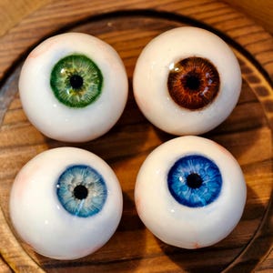 Op de afbeelding: Vier realistisch ogende glazen oogbollen met verschillende iriskleuren: groen, bruin, blauw en lichtblauw. De oogbollen liggen op een houten oppervlak.