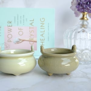 Puede incluir: Dos quemadores de incienso de cerámica, uno en un esmalte amarillo pálido y el otro en un esmalte verde apagado. Ambos tienen una forma redondeada con tres patas y dos asas.