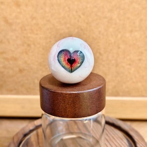 Op de afbeelding: Een gedetailleerd, handgemaakt glazen oog met een hartvormige iris. Het oog heeft een witte sclera met rode aderen, een veelkleurige iris en een zwarte pupil. Het rust op een houten basis boven een glazen pot, met een uniek ontwerp.