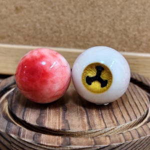 Op de afbeelding: Twee bolvormige oogbollen. Eén is effen rood met zichtbare aderen. De andere is wit met een gele iris en een zwarte pupil. De oogbollen worden op een houten oppervlak weergegeven.