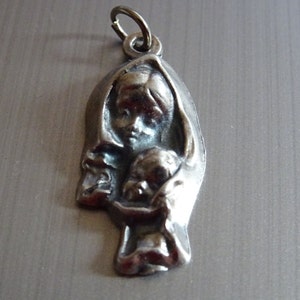 May include: Silver pendant featuring a detailed relief sculpture of the Virgin Mary holding a baby Jesus. The pendant has a loop at the top for attaching to a chain.