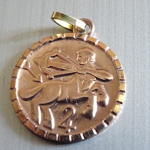 Pode incluir: Pingente dourado com uma representação detalhada do signo do zodíaco Sagitário. O pingente é redondo e tem uma borda texturizada.